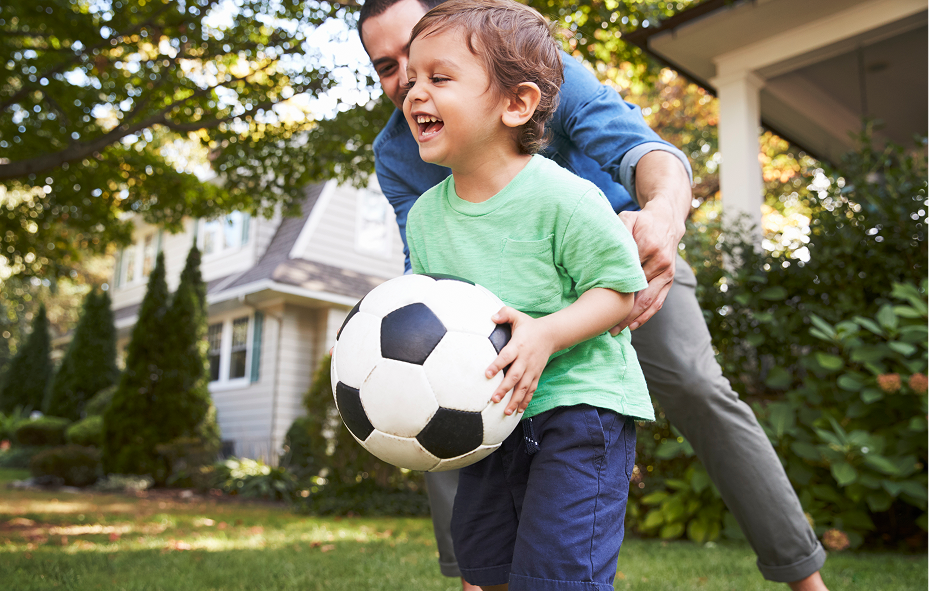 Young boy laughing and playing soccer with his father on a green lawn in front of a house