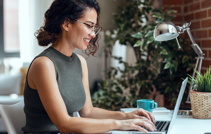 miling woman with glasses working on a laptop at a bright home office desk with plants and a coffee mug.